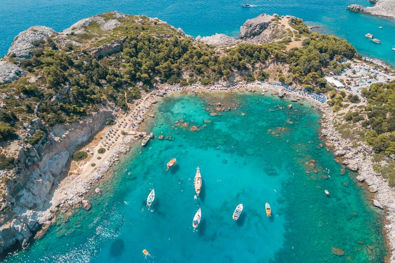 Aerial view of Anthony Quinn Bay with turquoise water and rocky, pine-covered shores